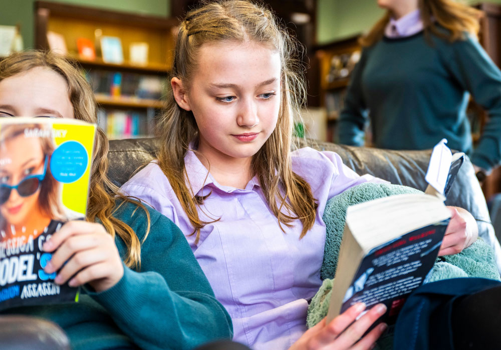 Senior student at Farnborough Hill School reading a book with friends in the library.